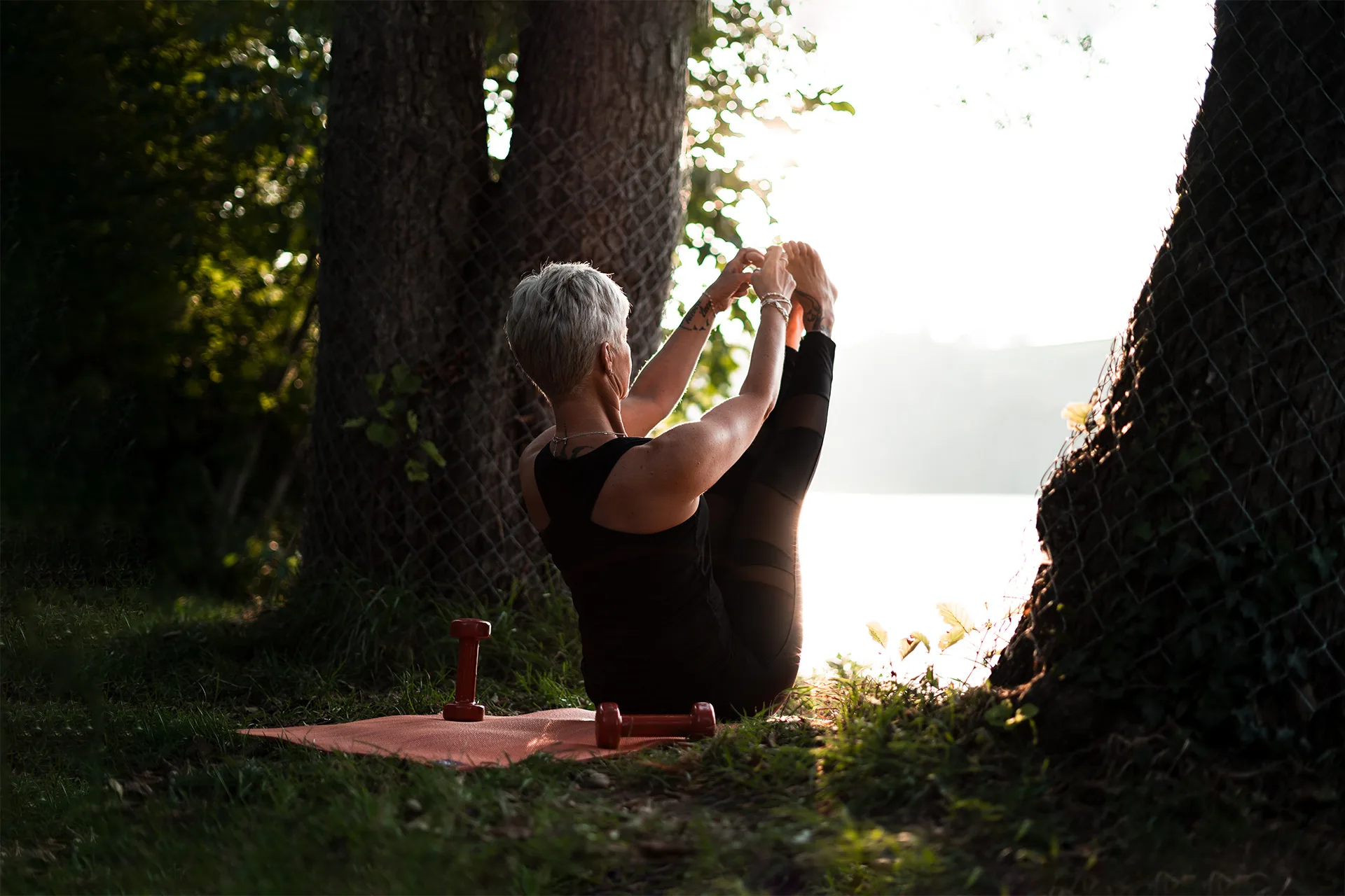 The trainer performs a seated stretch by a sunlit lake, illustrating the mindful, holistic approach to movement and wellbeing at FIT-N-FUN.
